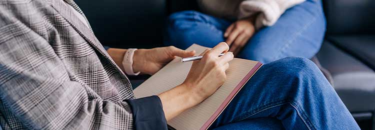 hands writing in notebook with person sitting in the background